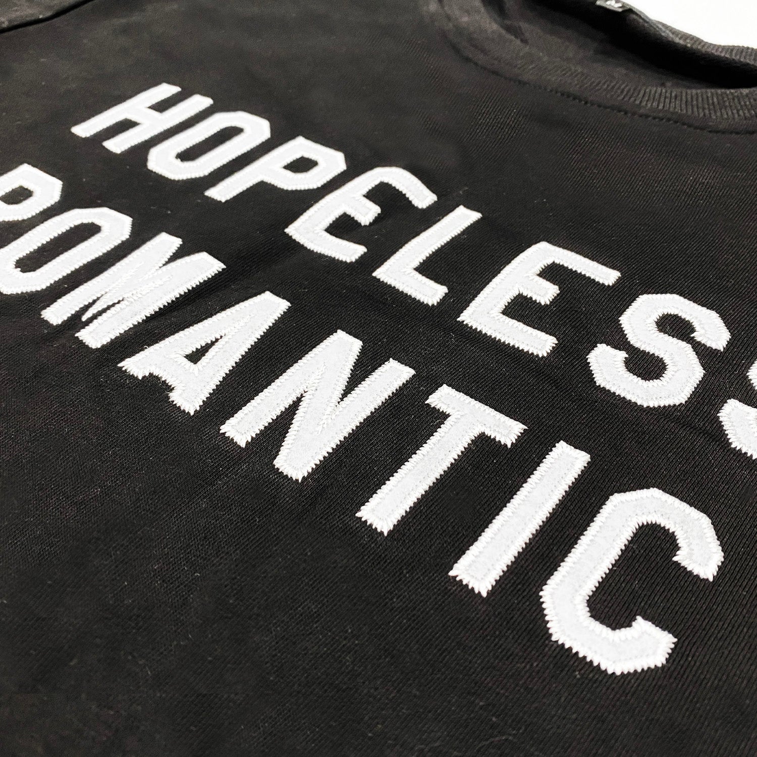  Close up image of a black crewneck sweatshirt with white felt letters that say hopeless romantic.